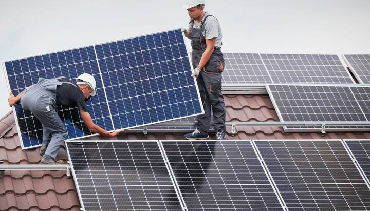 An image of a solar panel installation on the roof of a commercial building in Modesto, CA. The installation team can be seen working on the roof, positioning the panels to capture maximum sunlight. The city skyline can be seen in the background, with clear blue skies and a few clouds. Tools, equipment, and safety gear are scattered on the roof.